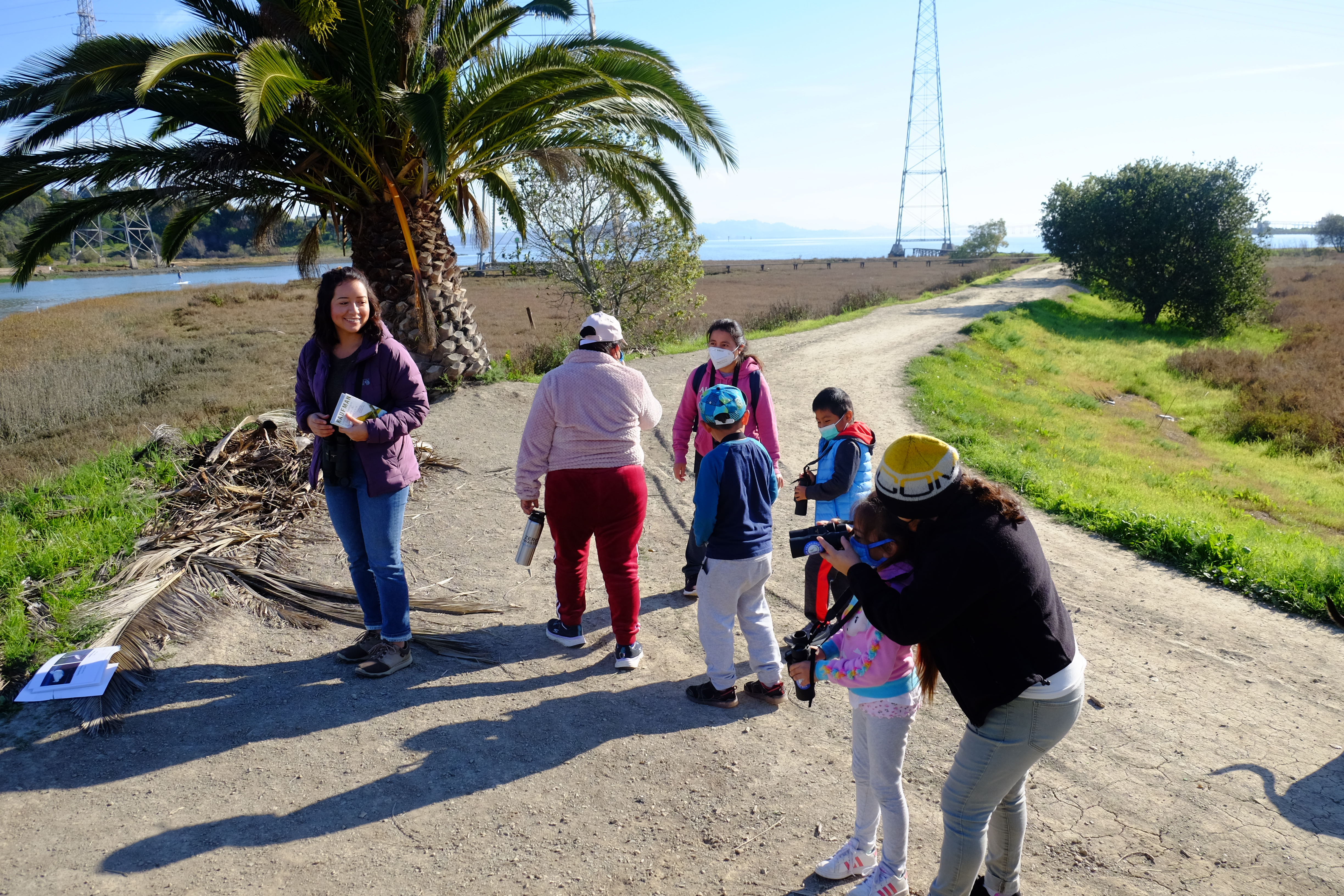 Education coordinator smiling and looking back at a group of mothers and children learning to use binoculars to look at birds. 