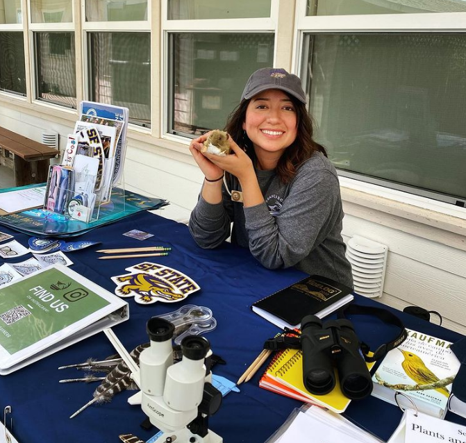 Picture of Bella wearing an SFSU hat holding up a stuffed animal salt marsh harvest mouse sitting at a table spread with science equipment demos, flyers, and stickers. 