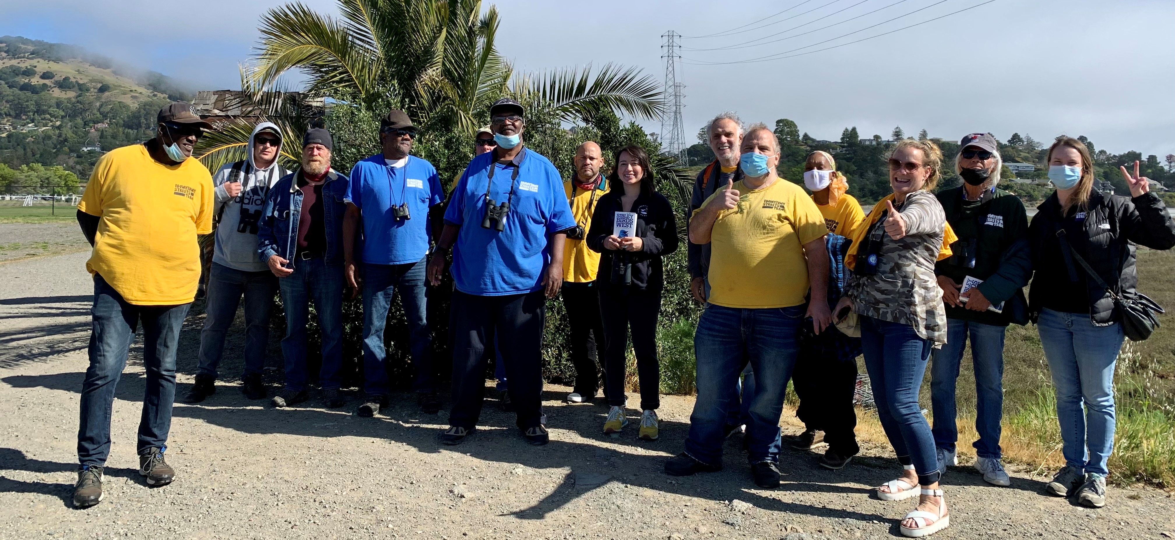 Group photo of Bella and Downtown Streets Team Members wearing bright blue and yellow tshirts smiling and standing in front of the marsh at Pickleweed Park.