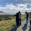 Group of people, some wearing shirts that say Downtown Streets Team, stand by the marsh overlooking the water with binoculars in hand to see birds.