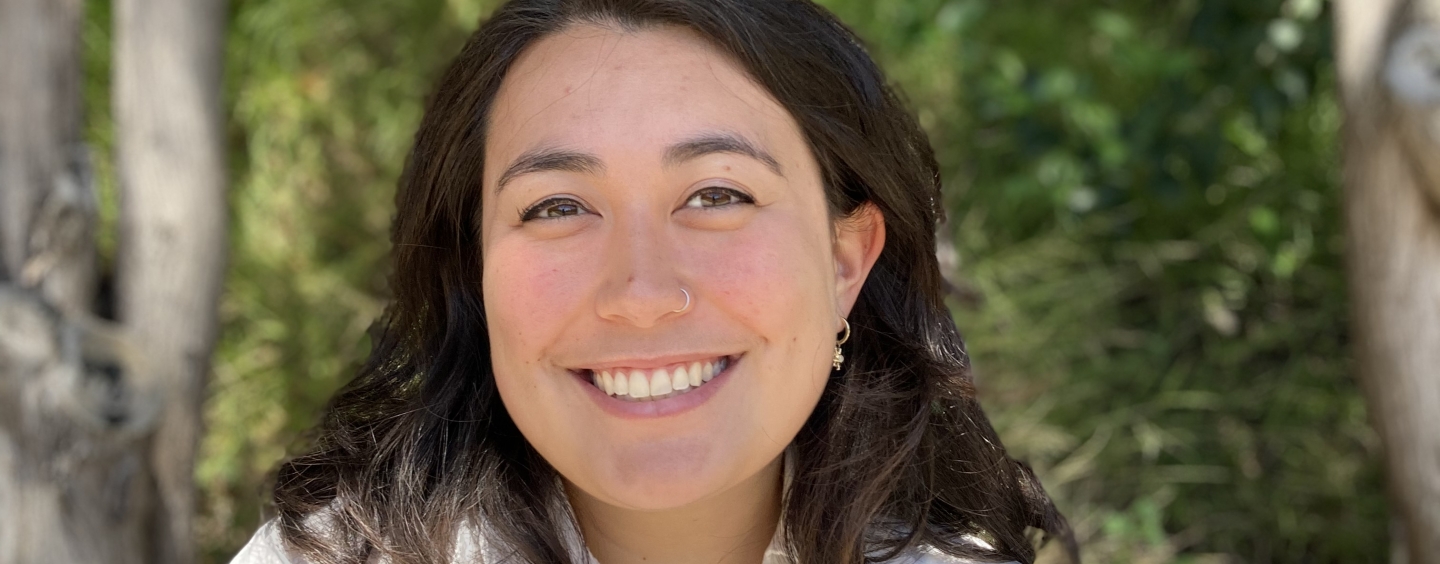 Headshot of Olivia Won smiling against background of trees and leaves. She has dark brown hair and is wearing a white linen shirt.
