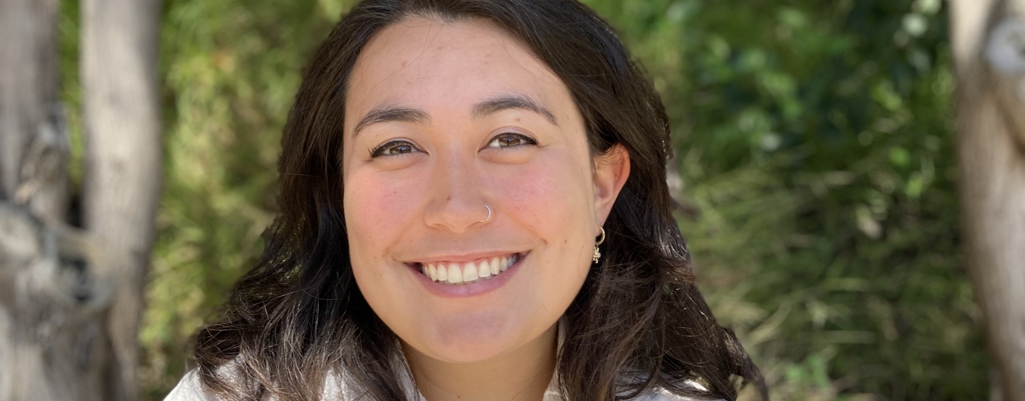 Headshot of Olivia Won smiling against background of trees and leaves. She has dark  brown hair and is wearing a white linen shirt.