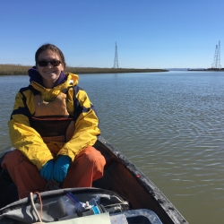 Picture of Danielle sitting in a kayak on smooth bay waters. She is smiling and wearing sunglasses wearing a bright yellow raincoat and a life vest. 