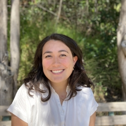 Headshot of Olivia Won smiling against background of trees and leaves. She has dark  brown hair and is wearing a white linen shirt.