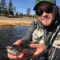 Jeremiah wearing camo, sunglasses, and a hat holding a fish. He is standing out over a body of water with trees in the background.