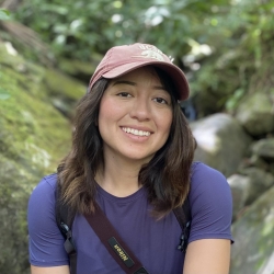 Picture of Bella with short brown hair and a red cap smiling in front of a nature background.